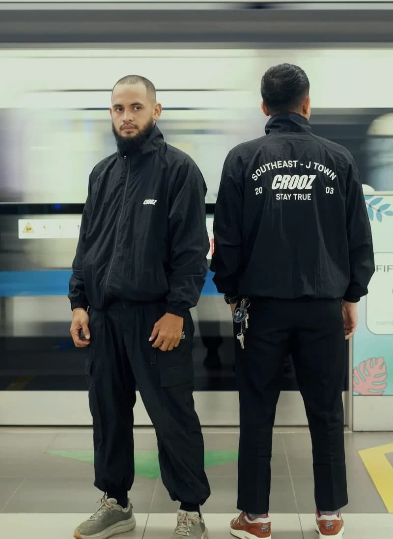 Dua model di peron MRT memakai jaket nylon hitam, satu menghadap depan dengan logo di dada, satu membelakangi dengan sablon punggung