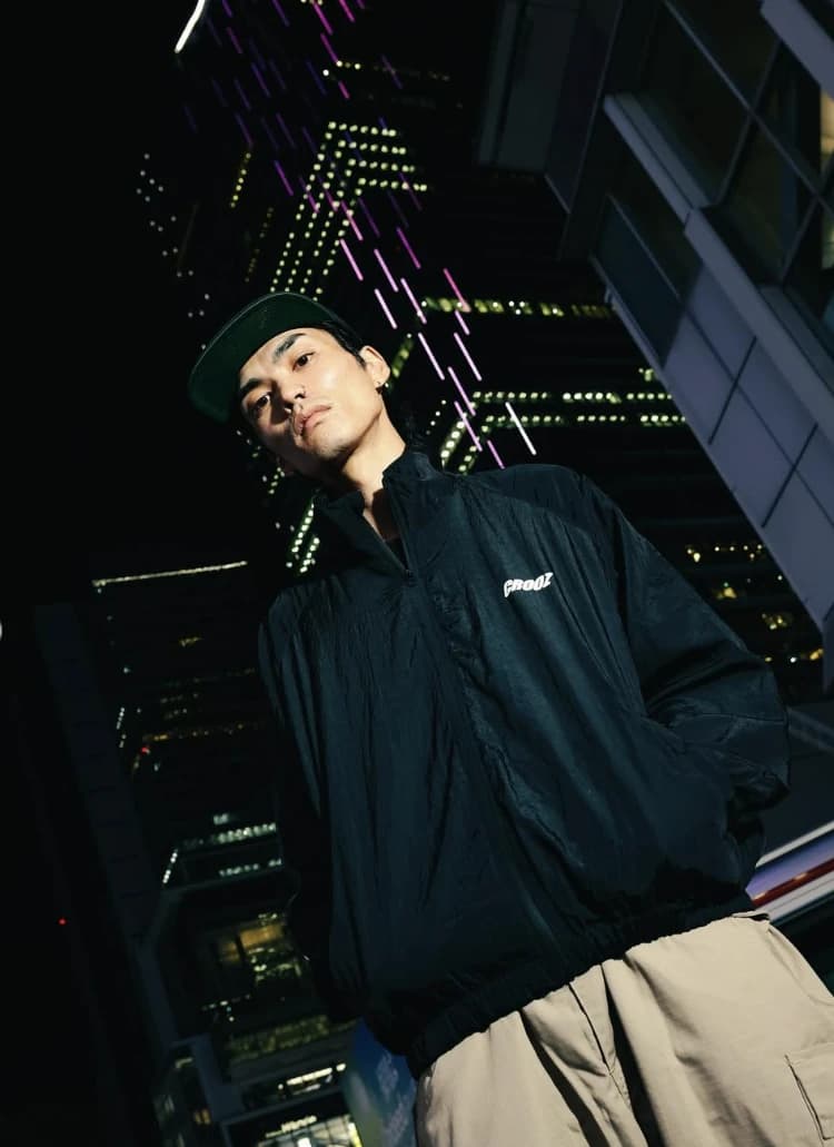 Model memakai jaket nylon hitam berlogo CROOZ di dada, difoto dari bawah dengan gedung menyala di malam hari