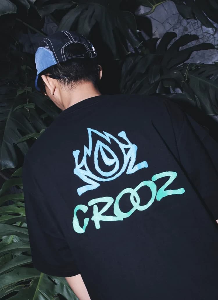 Punggung kaos hitam dengan ikon flame dan tulisan CROOZ gaya graffiti bergradasi biru hijau, berlatar daun monstera