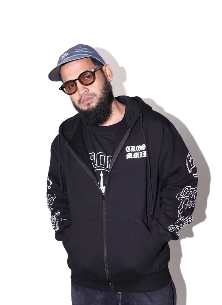 Model memakai MMIII Zip Hoodie hitam dengan sablon lengan putih, di depan dinding studio putih