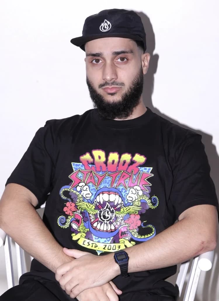 Model duduk memakai kaos hitam bergrafis monster warna-warni bertuliskan Crooz Stay True Estd. 2003
